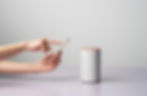 Hands holding a smartphone point at a gray smart speaker with an orange top. The setting is a light gray background on a purple surface.