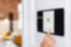 Finger taps a security app on a smart home screen. White wall background, blurred room view with yellow couch. Calm and modern atmosphere.