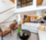 Modern living room with white sofa, brown chairs, and indoor plants. Wooden staircase and kitchen in the background; bright and airy.