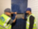 Two construction workers in safety vests from the company, Built to Perfection inspect blue herringbone tile in a partially completed room, ensuring it's built to perfection.