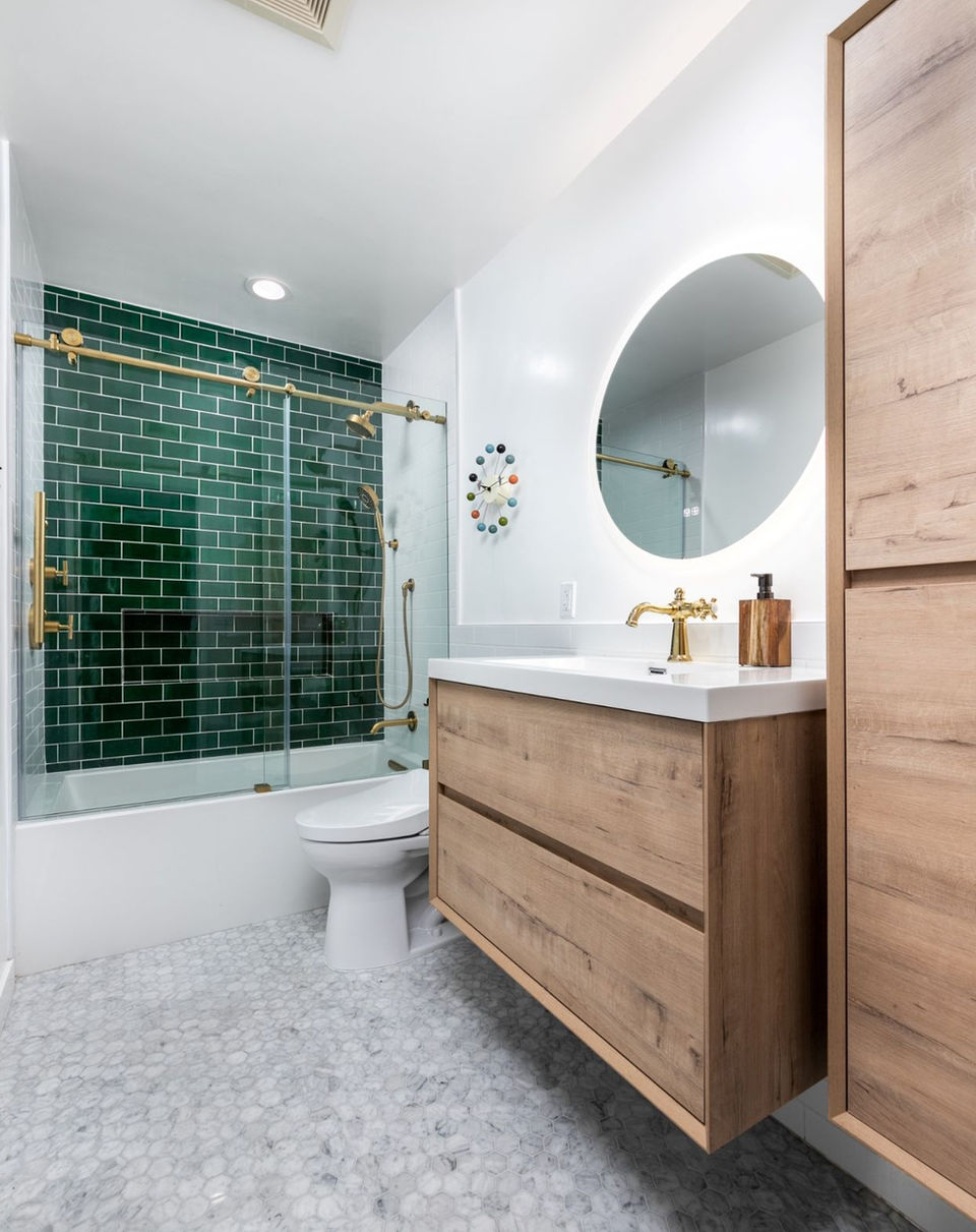 Modern bathroom with wooden vanity, round mirror, green tiled shower, and marble floor.