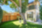 A lush backyard with a hammock hanging from a large tree. Modern house with sliding doors, wooden fence, green grass, and potted plants.