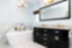 Modern bathroom with a white freestanding tub, black vanity with dual sinks, marble backsplash, gold fixtures, and soft lighting.