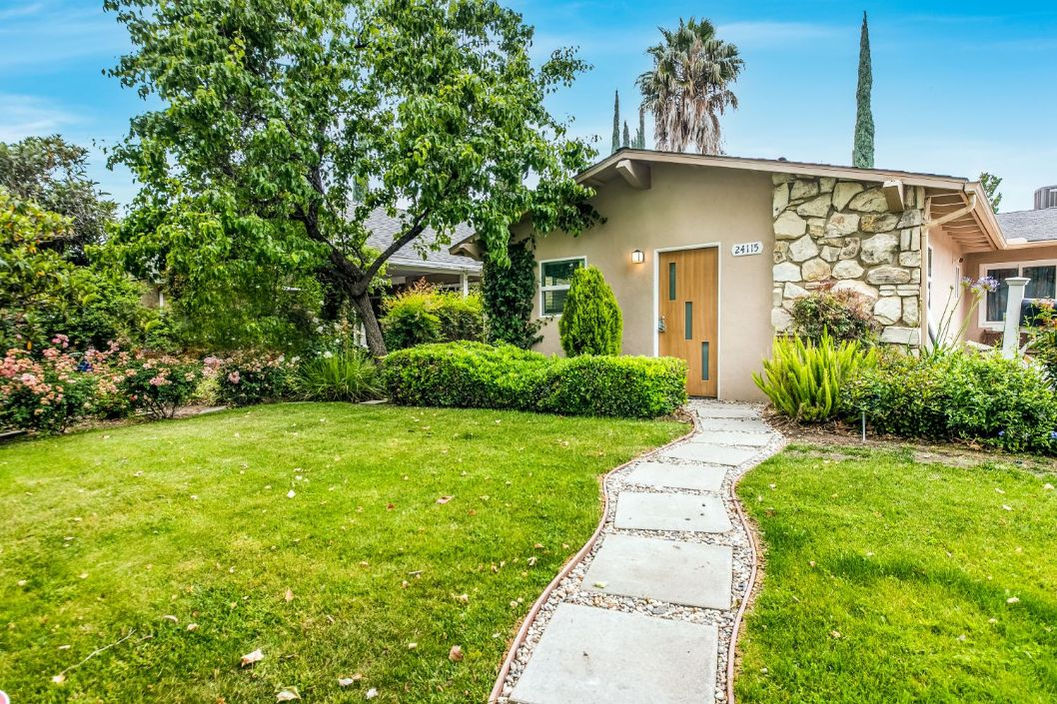 A small house with a stone facade, surrounded by greenery and a pathway leading through a grassy lawn.