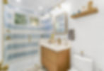 Bathroom with light blue striped tile shower, wooden vanity, and round mirror. Yellow flowers in vase, white walls, shelves with decor.
