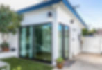 Modern white building with glass sliding doors, green potted plants, and a blue roof. Bright, sunny backyard setting with a white fence.