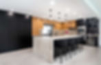 Modern kitchen with black and wood cabinets, marble island, and black chairs. Pendant lights hang above. Bright, sleek, and elegant.