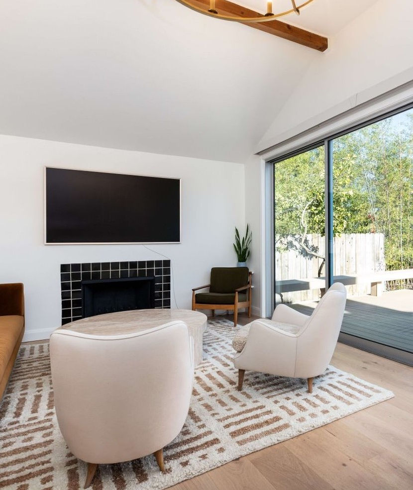 Modern living room with white armchairs, a fireplace, large TV, and floor-to-ceiling windows overlooking a garden.