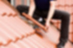 Person installing terracotta roofing tiles on a house roof. Wearing jeans and black shoes. Bright, clear sky in background.
