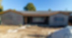 A house under construction with a partially built exterior, plastic-covered windows, and open door. Dirt and tools are visible in the yard.