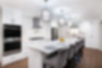 Modern kitchen with a long white island, gray chairs, stainless steel appliances, and pendant lights. Fruit bowl on counter, wood floor.