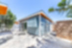 Modern blue house with sliding glass door, trees, and stone patio. Bright, sunny day with clear skies and shadows on the ground.