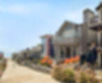 Beachfront houses with balconies overlook a path to the sea. Colorful foliage lines the walkway under a clear blue sky. Peaceful scene.