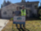 Two men stand smiling in front of a house under renovation, holding a sign from general contractor, Built to Perfection.