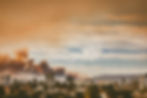 A city skyline with palm trees under a smoky sky. Dark smoke billows from a building, creating an ominous mood against orange clouds.